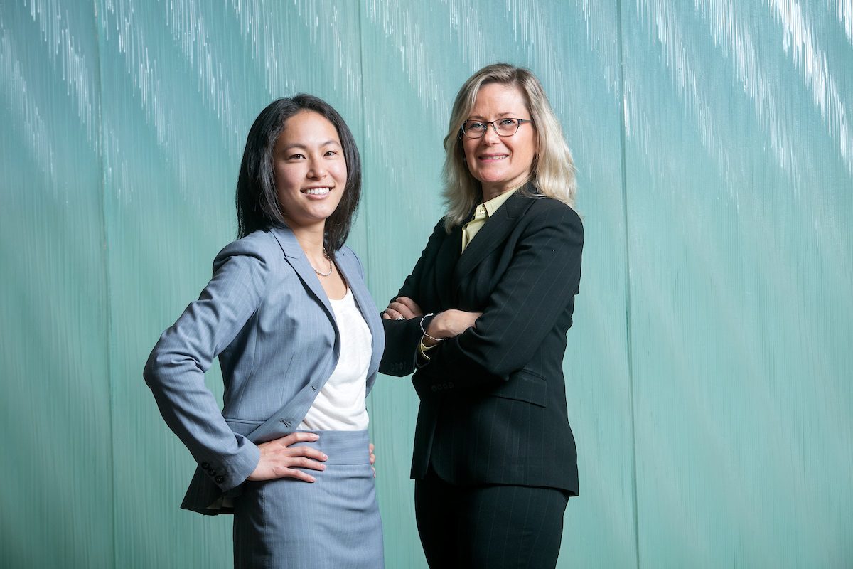 Two UC Law SF women posing for photo, standing facing each other, looking and smiling towards the camera. Woman on the left is in a gray suit jacket and white top. Woman on the left is wearing glasses and a black suit jacket and white top.