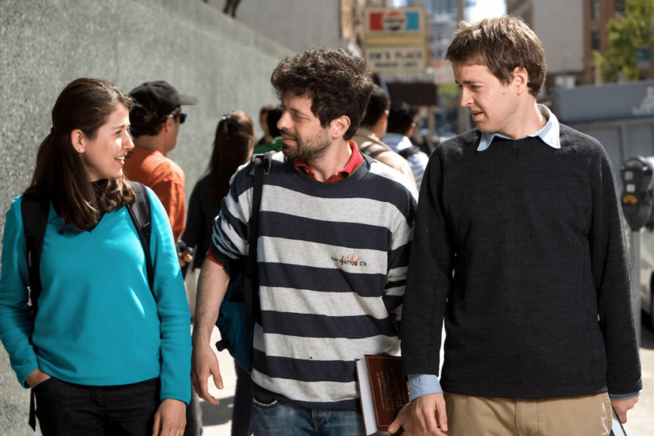 Students (woman in teal sweater on the left, man in striped black and white sweater in the middle and a man wearing a black sweater with white collars on the right) walking together outside a building.