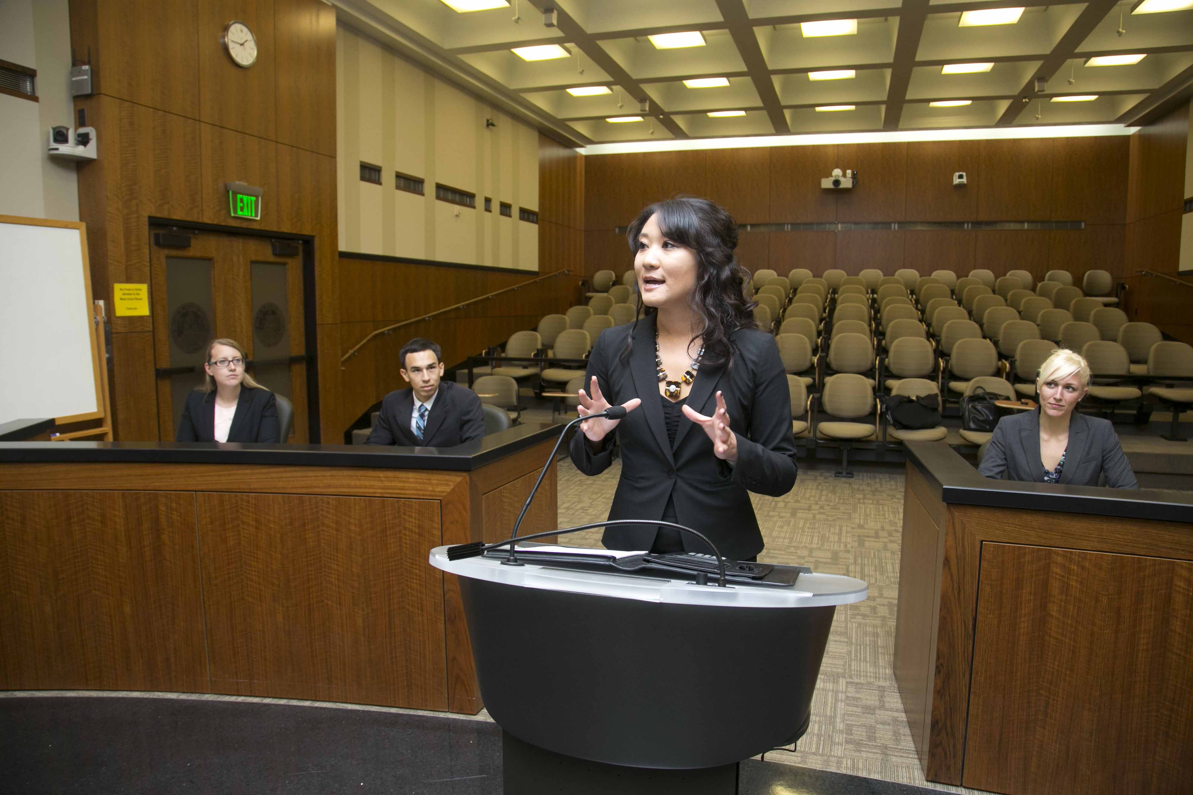 A student speaking in moot court, three individuals are sitting behind her