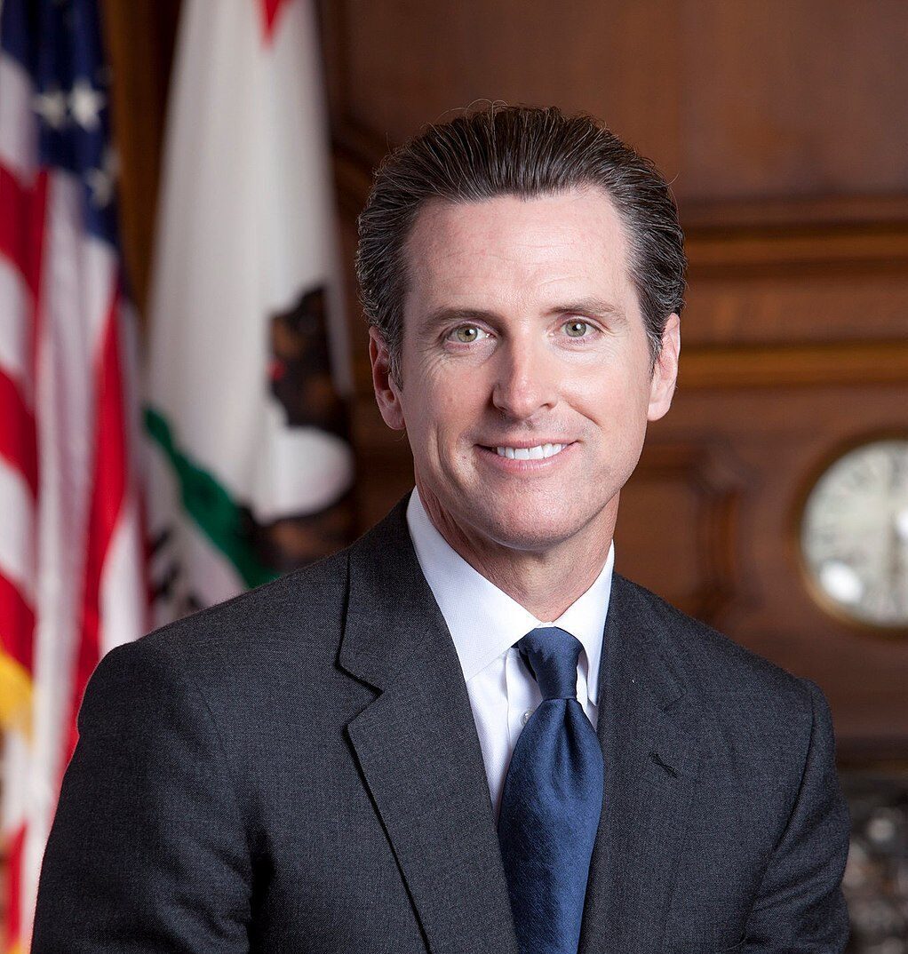Man smiling at the camera wearing a suit. Backdrop: United States flag and California state flag