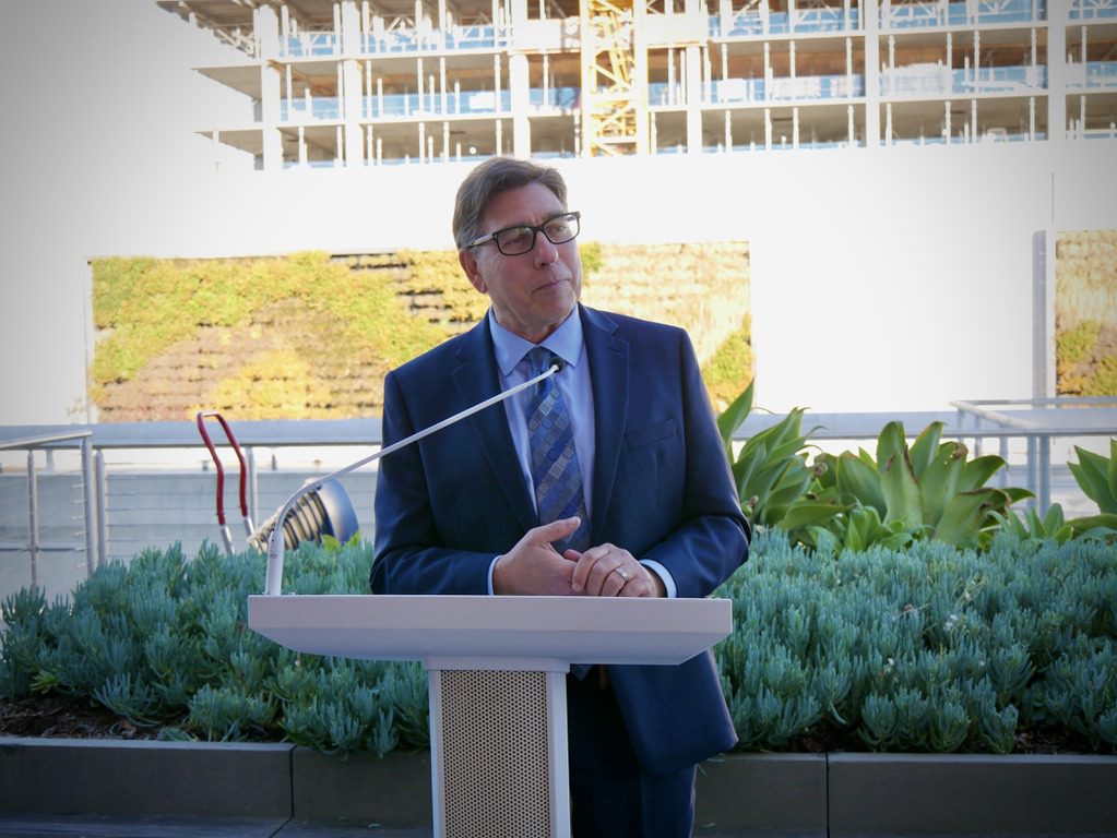 Man wearing glasses and a blue suit, talking at a podium with greenery and building construction in the backdrop